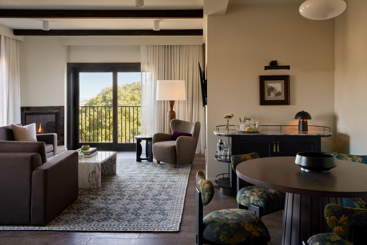 A modern living room with a fireplace, a dining area with a round table and floral chairs, and a view of the outdoors through large windows.