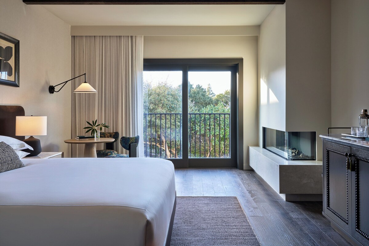A modern hotel room with a large bed, a desk with a lamp, a small table with plants, and a balcony with a view of trees.
