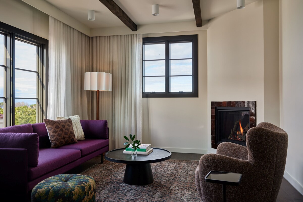 A modern living room with a purple sofa, a round coffee table, a patterned ottoman, a fireplace, and large windows with curtains.