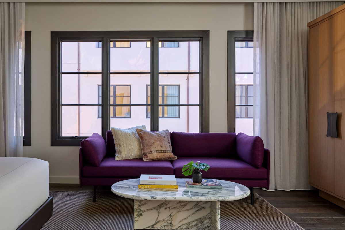 Ein Zimmer mit einem violetten Sofa, einem Marmor-Couchtisch und großen Fenstern mit Blick auf Gebäude.
