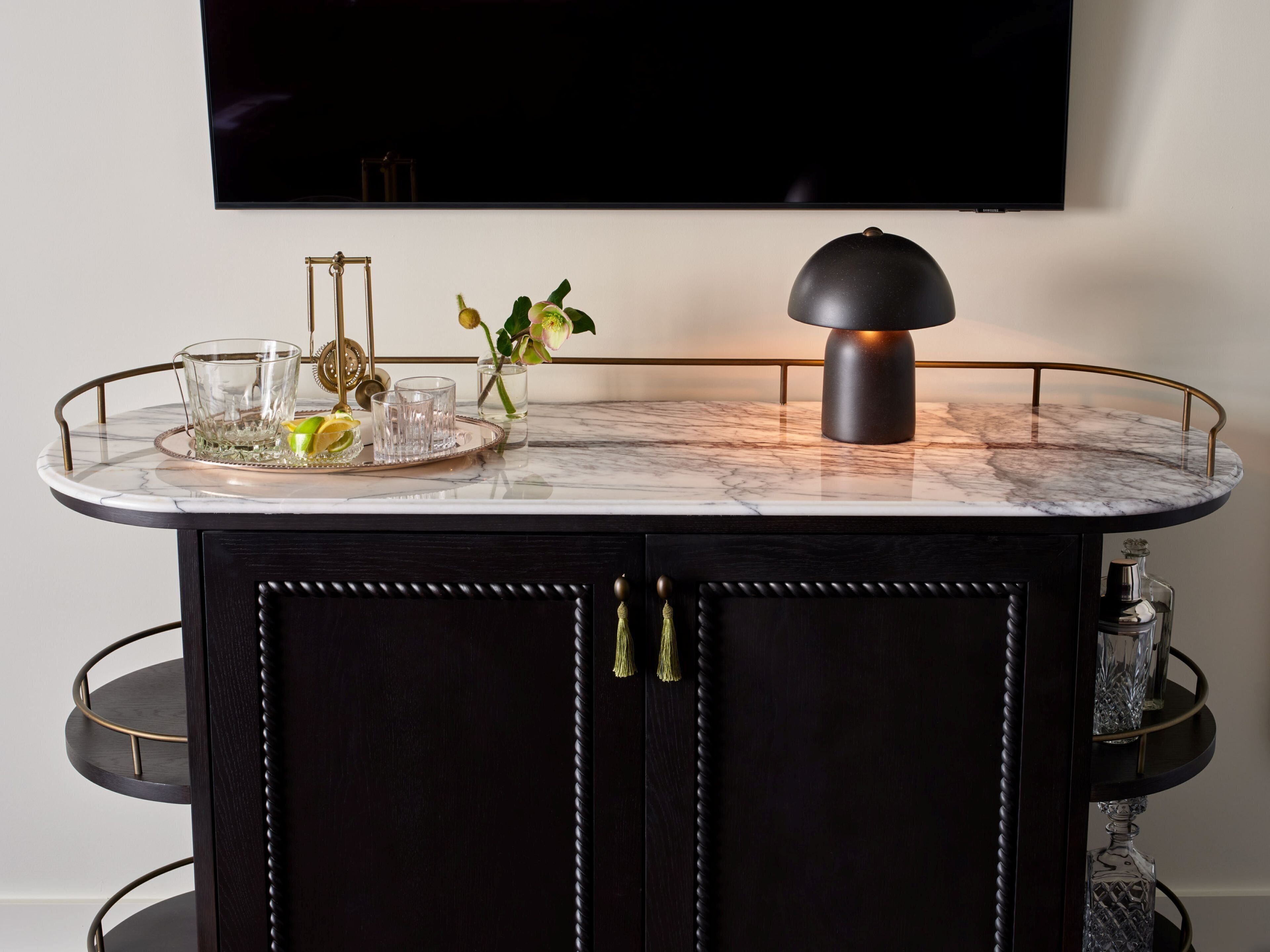 Un mueble de madera oscura con encimera de mármol, una bandeja de vidrio con un limón, vasos y un jarrón con flores.