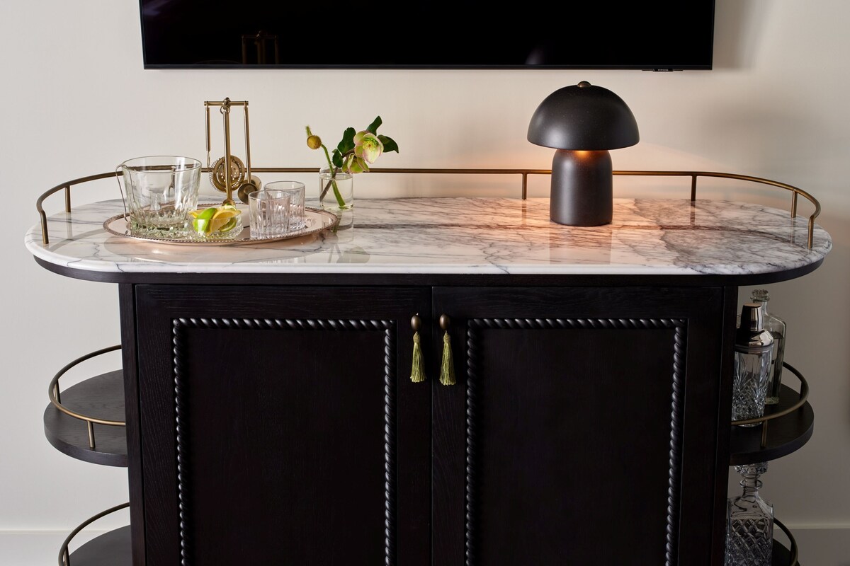 A dark wooden cabinet with a marble countertop, a glass tray with a lemon, glasses, and a flower vase.