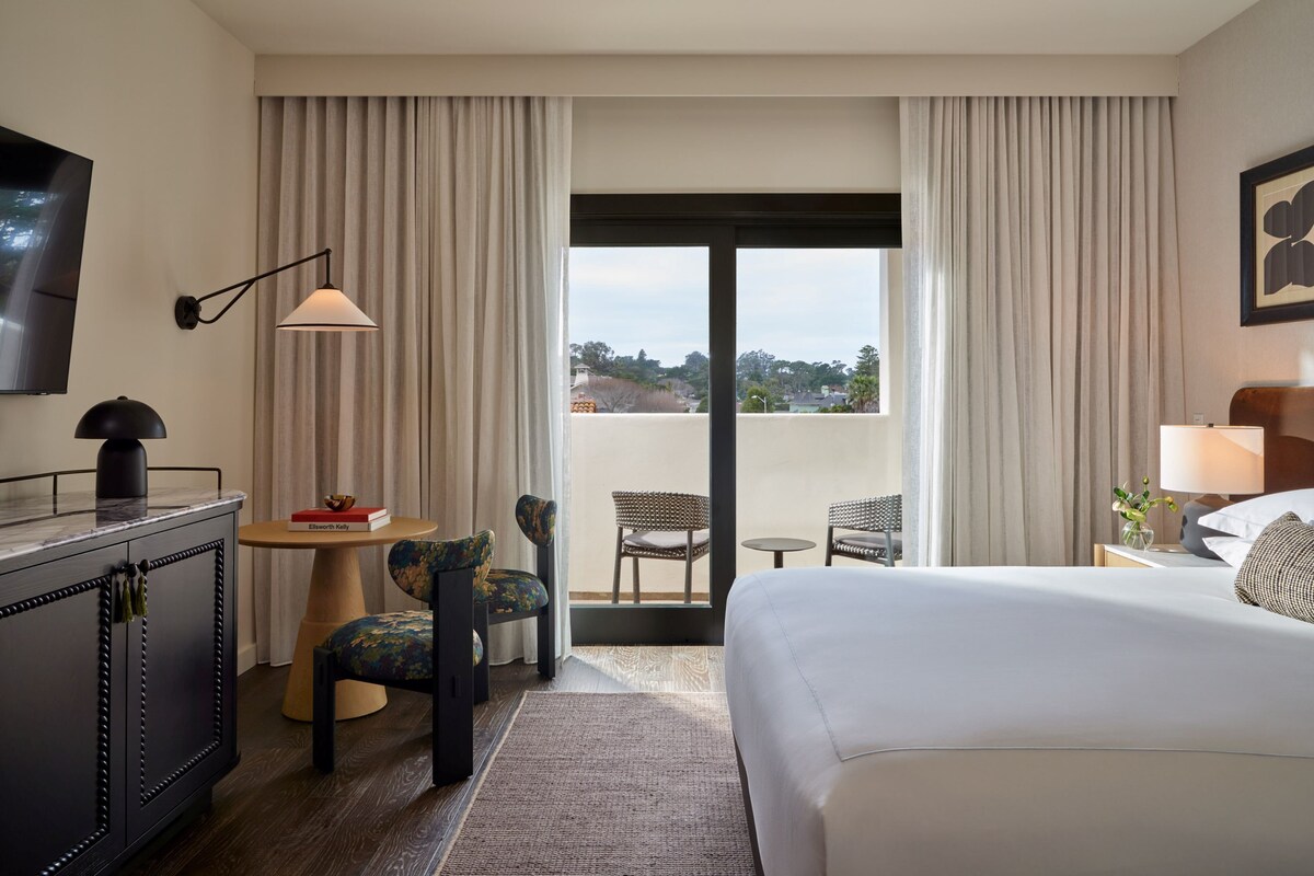 A hotel room with a bed, a desk with a lamp, a chair, a small table with books, and a view through a large window.