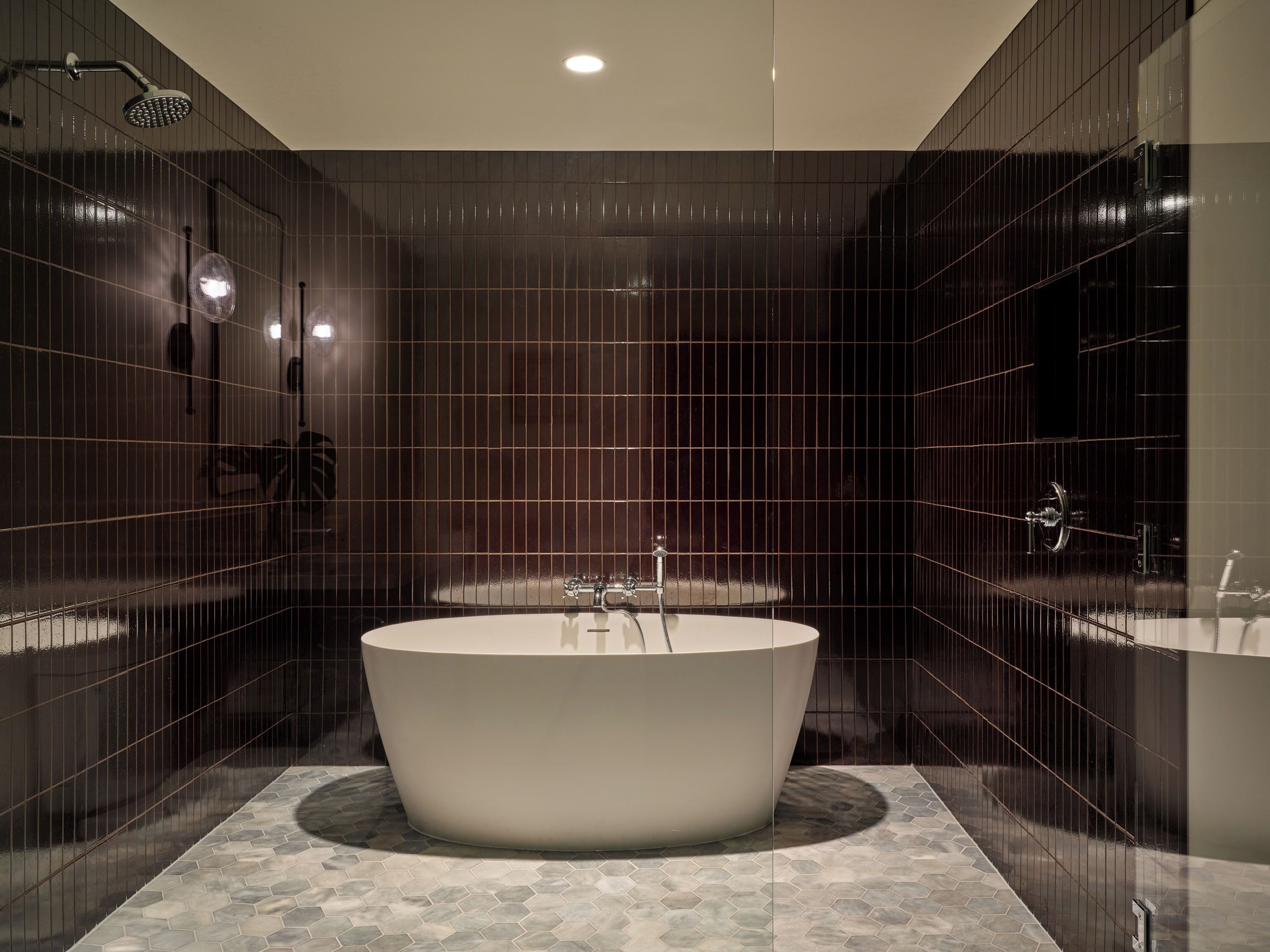 A modern bathroom with a freestanding bathtub, dark tiled walls, and a hexagonal tile floor.