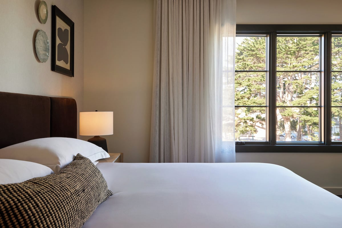A hotel room with a large bed, a nightstand with a lamp, a window with curtains, and wall art.