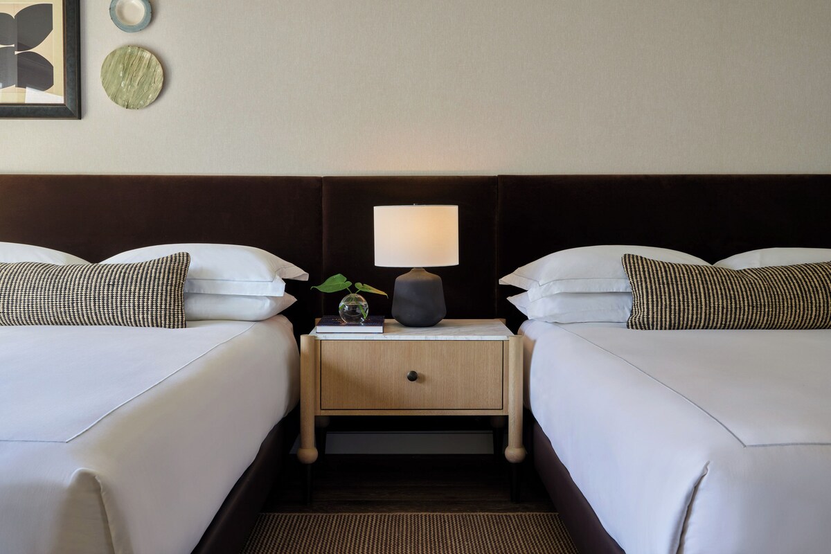 A hotel room with two beds, a nightstand with a lamp, and a small plant.