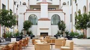 Un amplio espacio al aire libre con mobiliario de mimbre y sofás, una chimenea central y vistas a un edificio blanco con ventanas arqueadas.