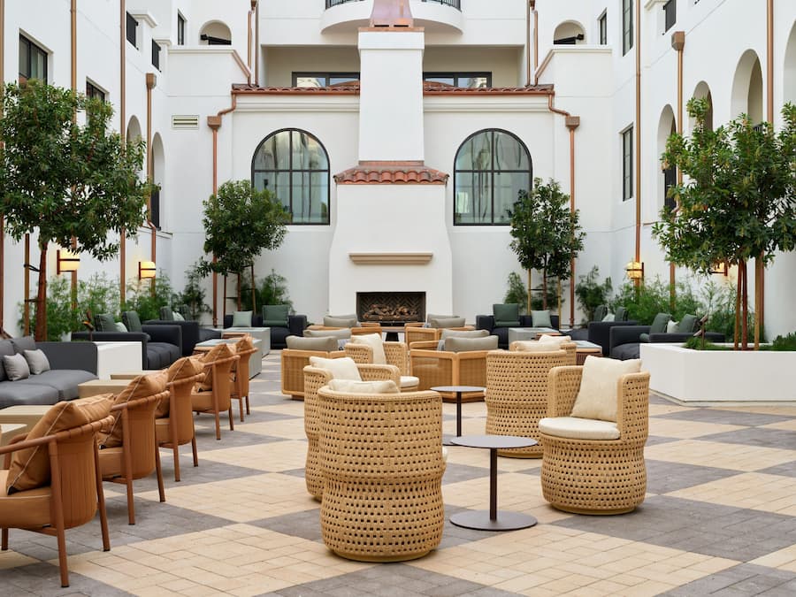 Un amplio espacio al aire libre con mobiliario de mimbre y sofás, una chimenea central y vistas a un edificio blanco con ventanas arqueadas.