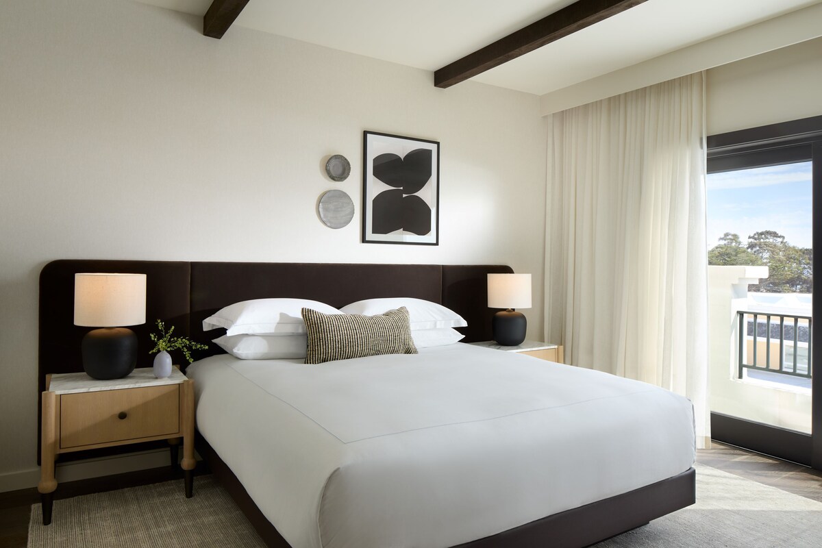 A modern hotel room with a large bed, two bedside tables with lamps, a view of the outdoors through a large window, and a framed artwork on the wall.