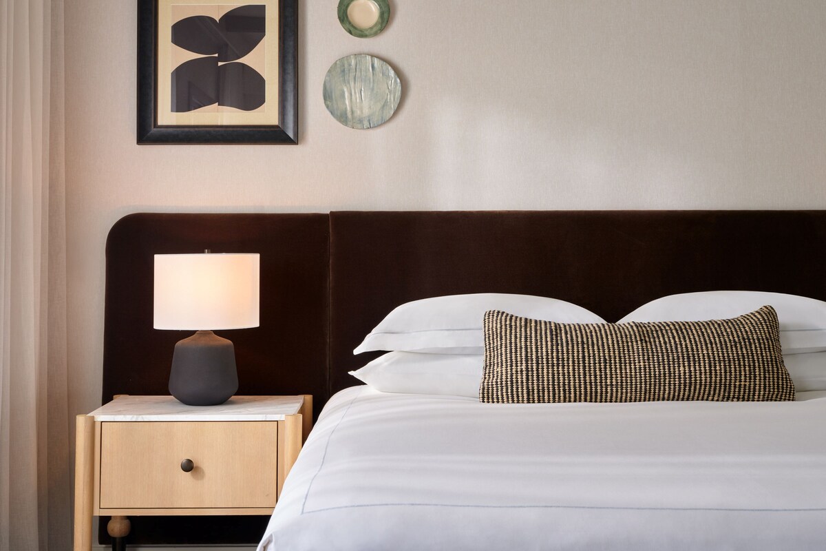 A neatly made bed with white linens and a patterned pillow, a bedside lamp on a wooden nightstand, and a framed artwork above the bed.