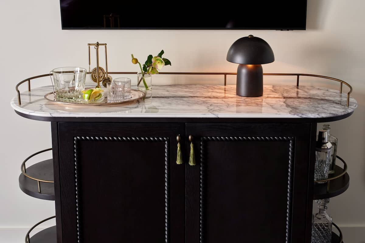 A dark wooden cabinet with a marble countertop, a glass tray with a lemon, glasses, and a flower vase.