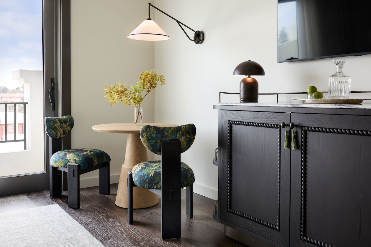 A modern living room with a dark wooden cabinet, a round table, two upholstered chairs, a floor lamp, and a television mounted on the wall.