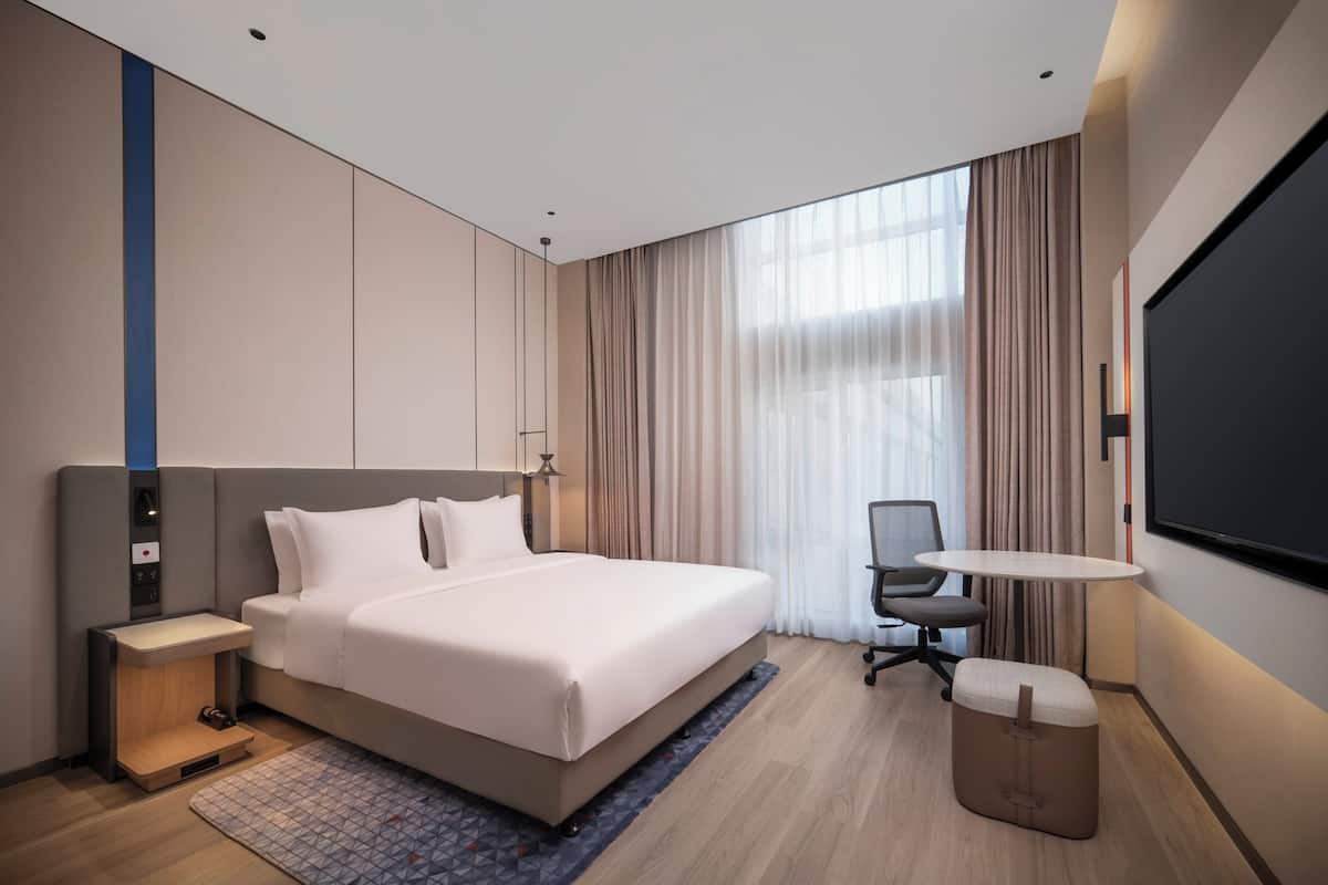 A modern hotel room with a large bed, bedside table, desk, chair, and a TV.