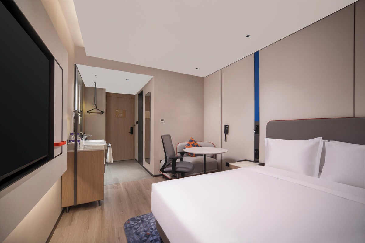 A modern hotel room with a bed, a desk, a chair, a small table, a mirror, a hanging light fixture, and a bathroom with a sink and mirror.
