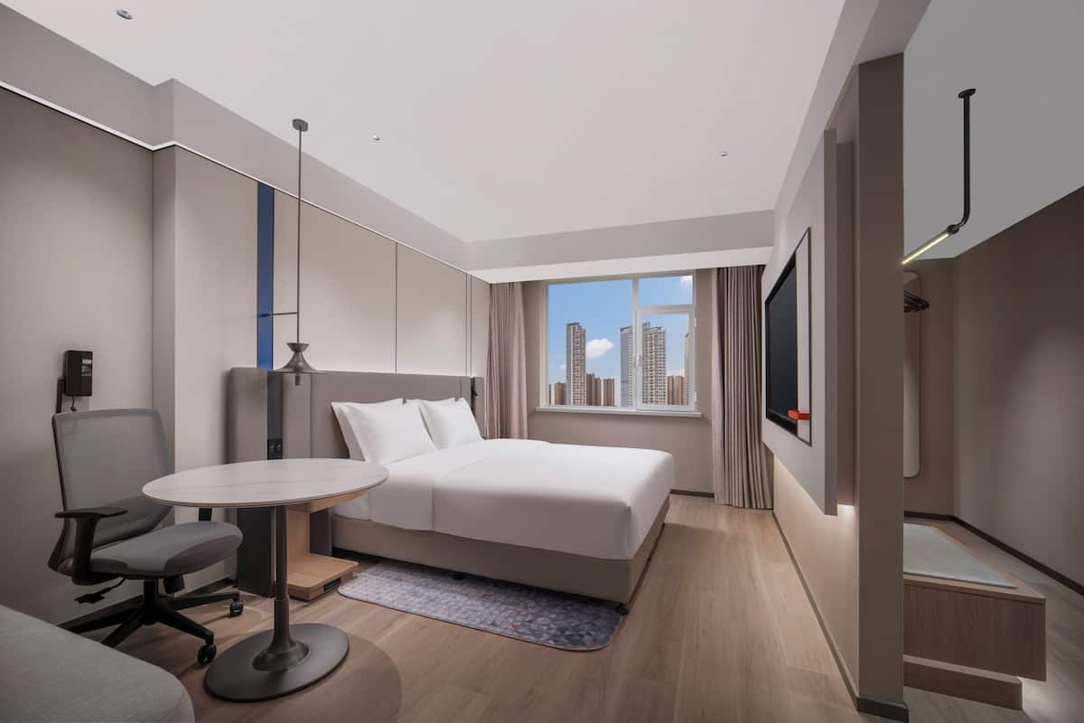 A modern hotel room with a large bed, a desk, a chair, and a view of the city skyline.