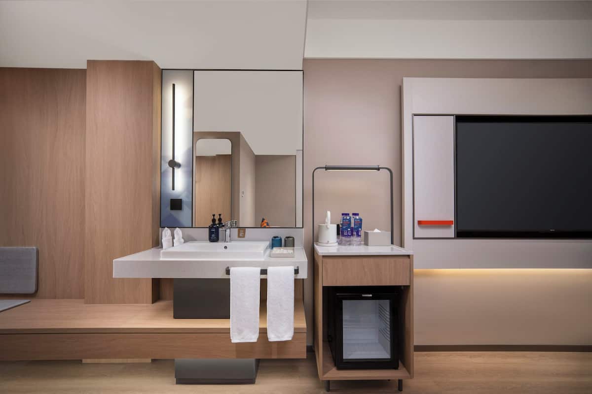 A modern bathroom with a large mirror, a sink with two faucets, a towel rack, and a small refrigerator.