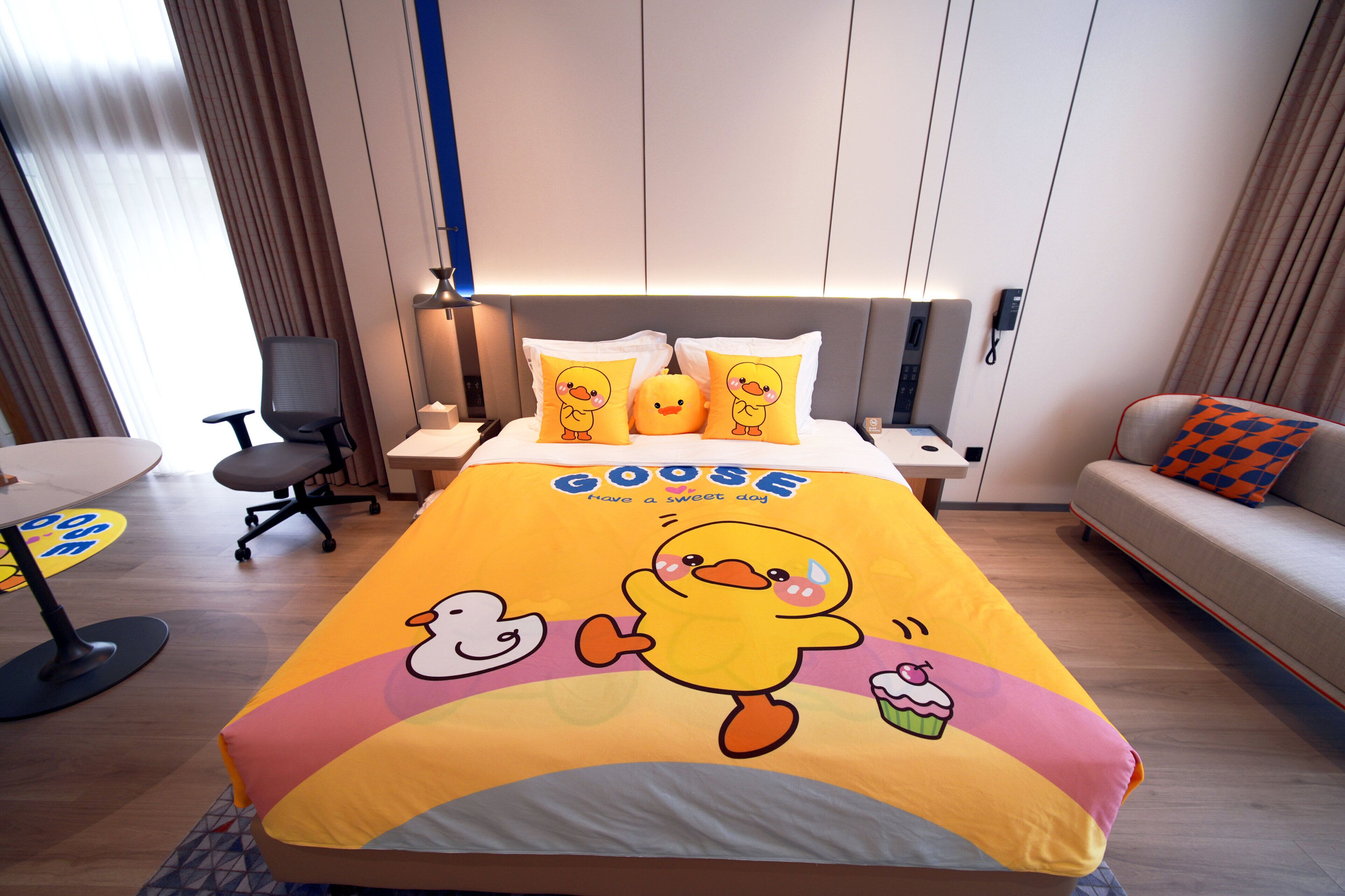Habitación de hotel con cama con edredón con estampado de patos, un escritorio con silla, un sofá y una mesita de noche con lámpara.