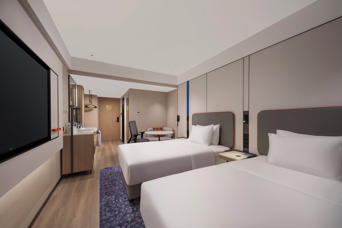 A hotel room with two beds, a desk with a chair, a small table, and a bathroom area.