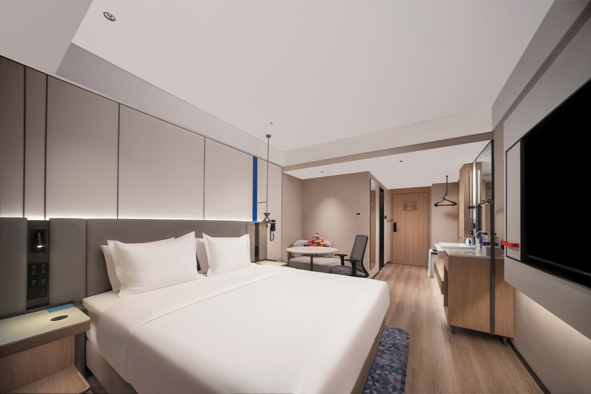 A modern hotel room with a large bed, bedside table, and a desk with a chair. There is a small table with a chair and a TV mounted on the wall.