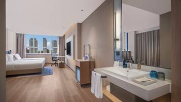 A modern hotel room with a large bed, a desk, a bathroom with a large mirror, and a view of tall buildings.