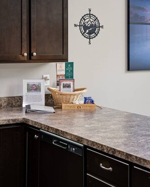 A kitchen with a granite countertop, a compass wall decoration, a wicker basket with a sign, and a framed welcome sign.