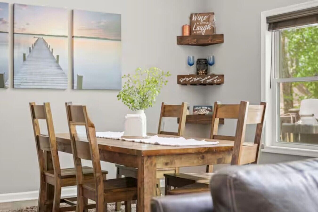 Un comedor con una mesa y sillas de madera, un jarrón con plantas verdes y decoraciones en la pared, incluyendo una pintura de un atardecer y un letrero que dice 'Wine a Little Laugh a Lot'.