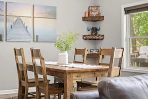 A dining area with a wooden table and chairs, a vase with greenery, and wall decorations including a sunset painting and a sign that says 'Wine a Little Laugh a Lot'.