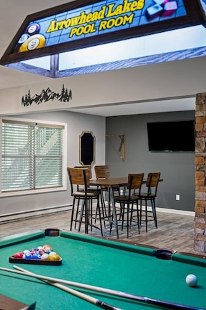 A pool room with a pool table, chairs, and a television screen displaying 'Arrowhead Lakes POOL ROOM'.
