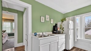 A kitchen with a white countertop, double sink, and a refrigerator. There are framed pictures on the wall and a plant on the counter.