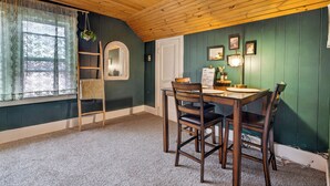 A dining area with a wooden table, chairs, a ladder, and a window with lace curtains.