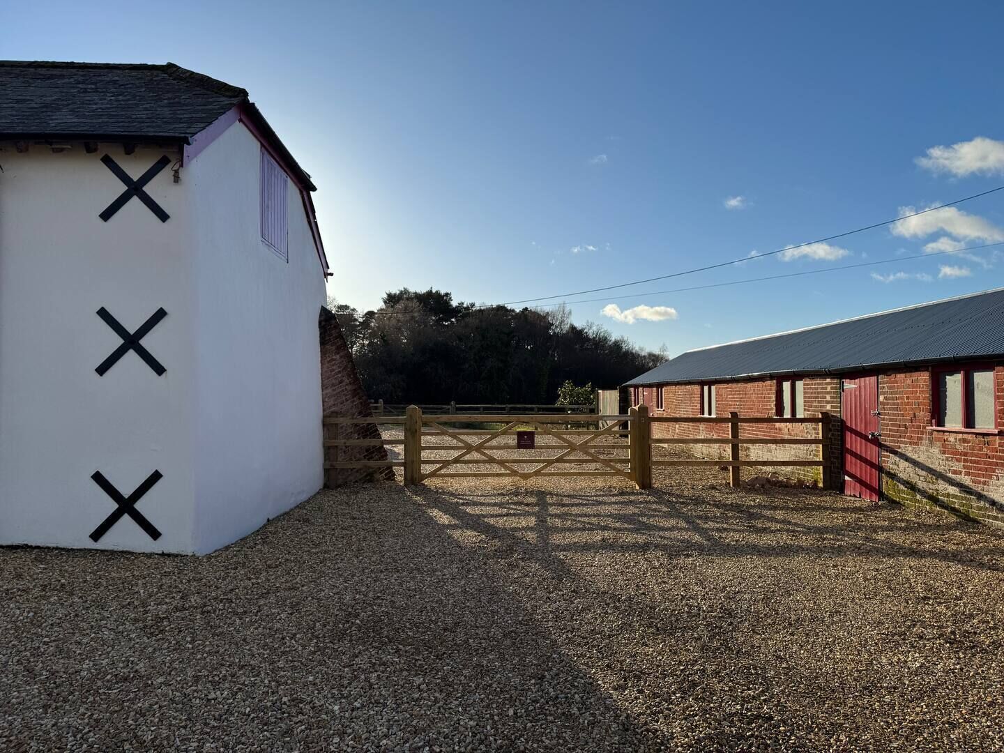 A gravel driveway leading to a gated area with a white building marked with black crosses, a red barn, and a clear sky.