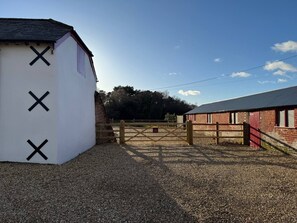 Une allée de gravier mène à une zone fermée par un portail, où se trouve un bâtiment blanc orné de croix noires, une grange rouge et un ciel dégagé.