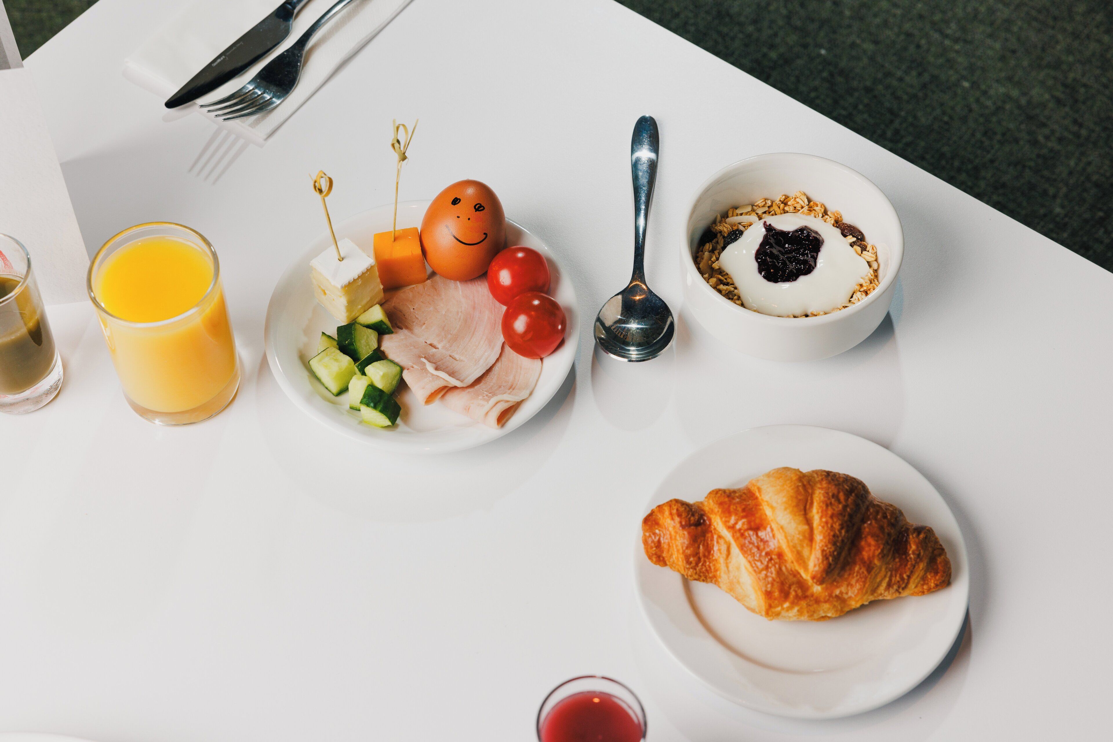 Un desayuno con un croissant, un vaso de zumo de naranja, un plato de huevos, queso y verduras, y un bol de yogur con granola.