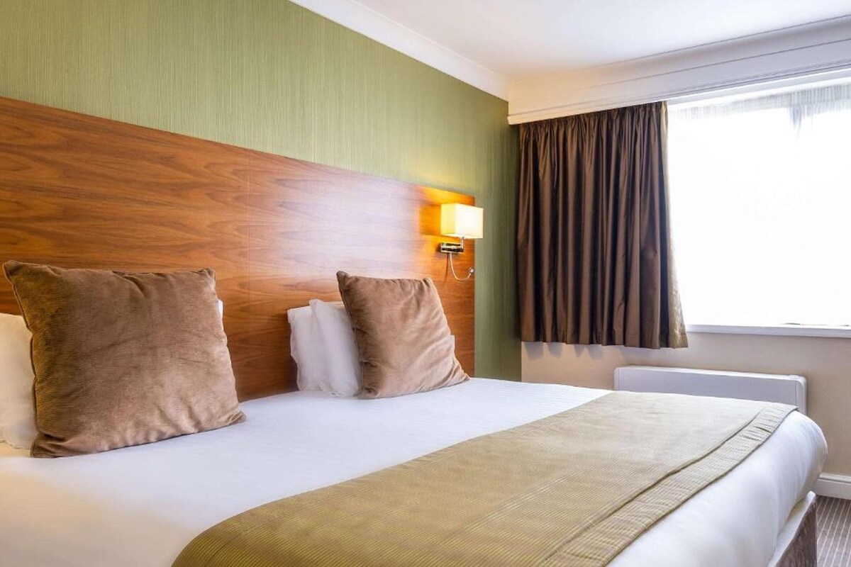 Une chambre d’hôtel avec un grand lit, deux oreillers, une lampe de chevet et une fenêtre avec des rideaux.
