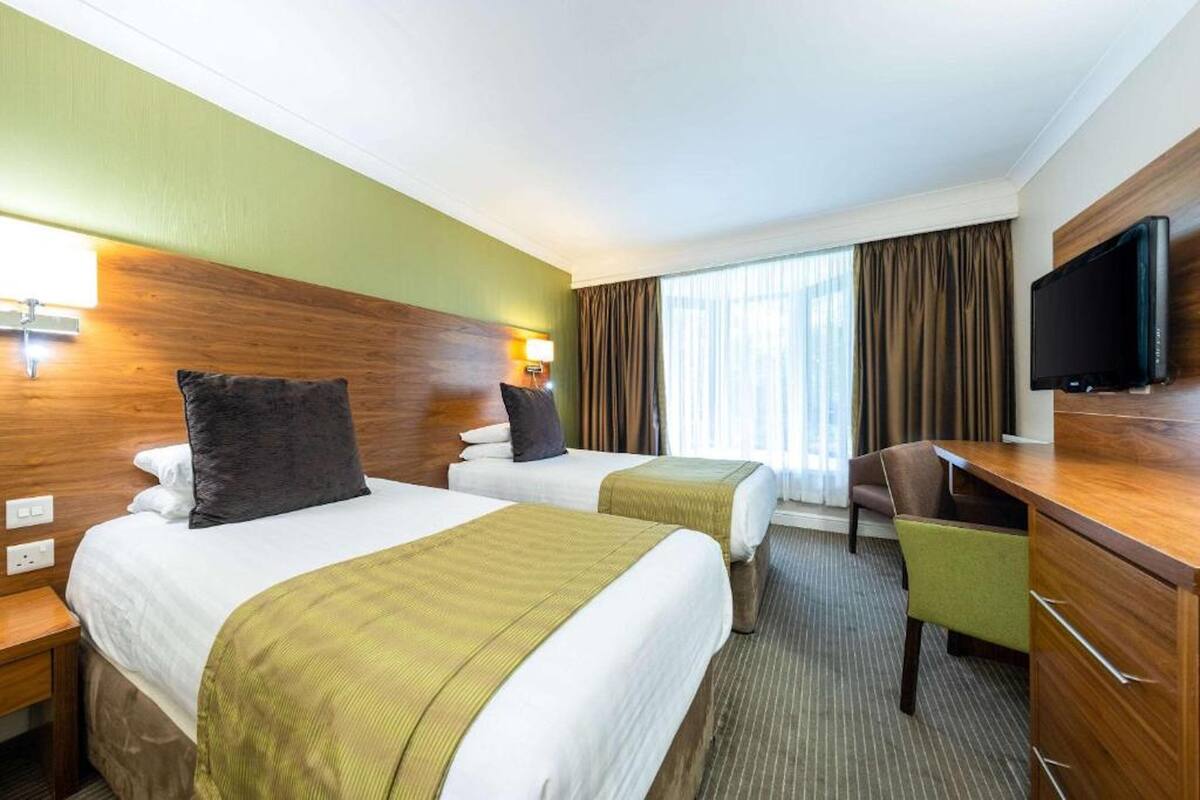 Ein Hotelzimmer mit zwei Betten, einem Schreibtisch, einem Stuhl, einem Fernseher und einem Fenster mit Vorhängen.