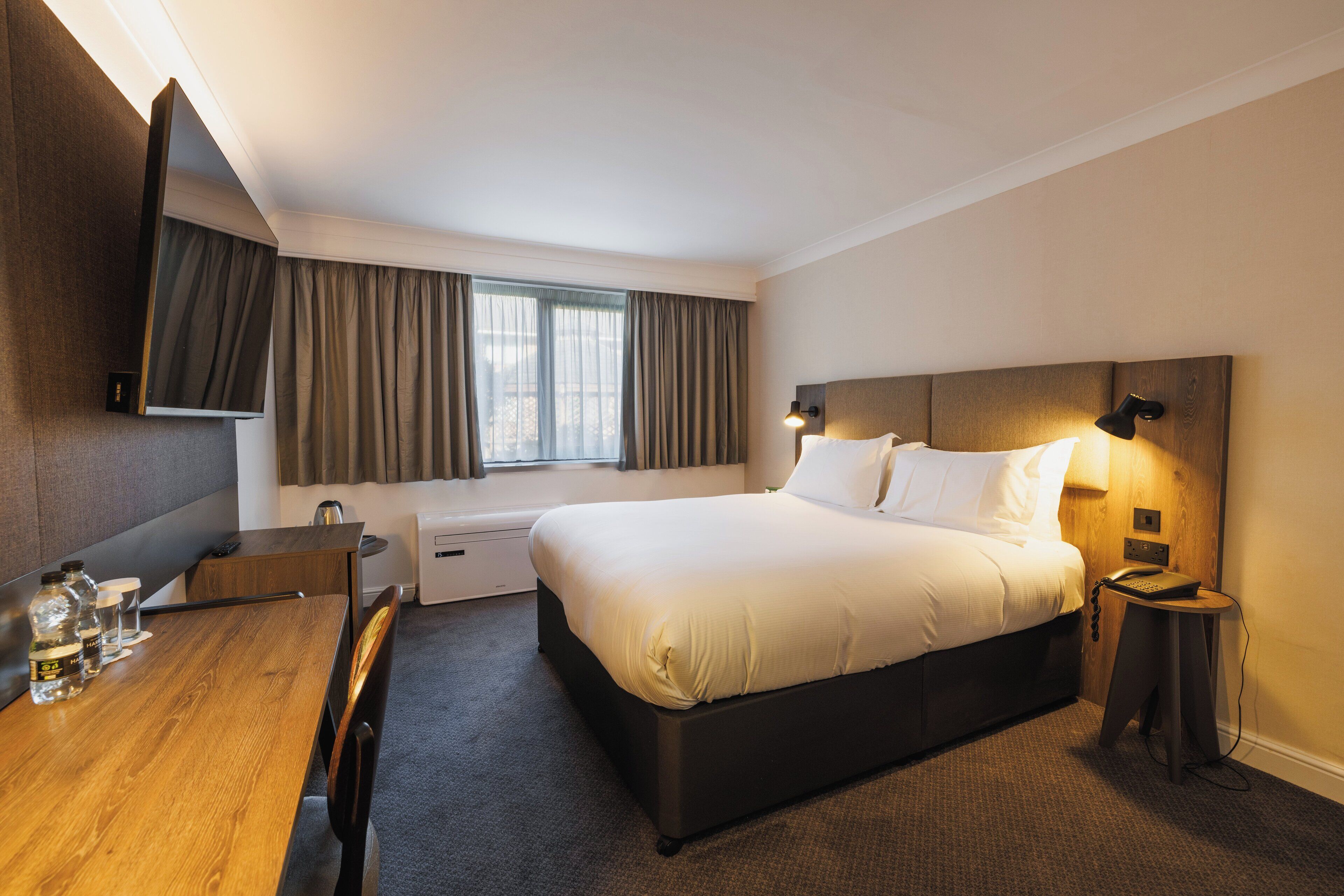 Ein Hotelzimmer mit einem großen Bett, einem Schreibtisch mit Stuhl, einem flachbildfernseher an der Wand, einem Nachttisch mit Lampe und einem Fenster mit Vorhängen.