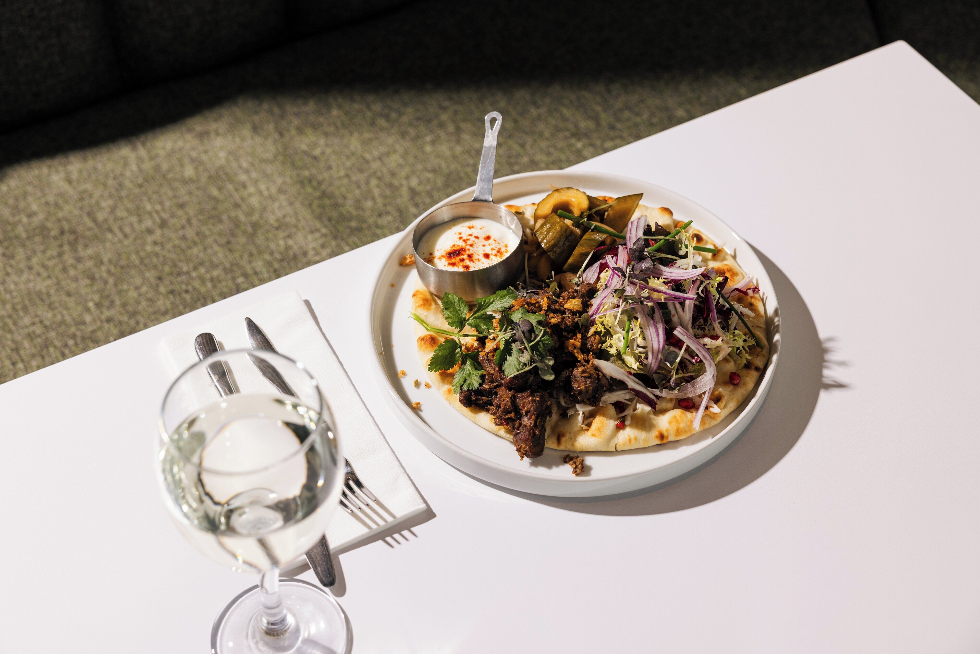 Een bord eten met een klein kommetje saus en een glas witte wijn op een witte tafel.