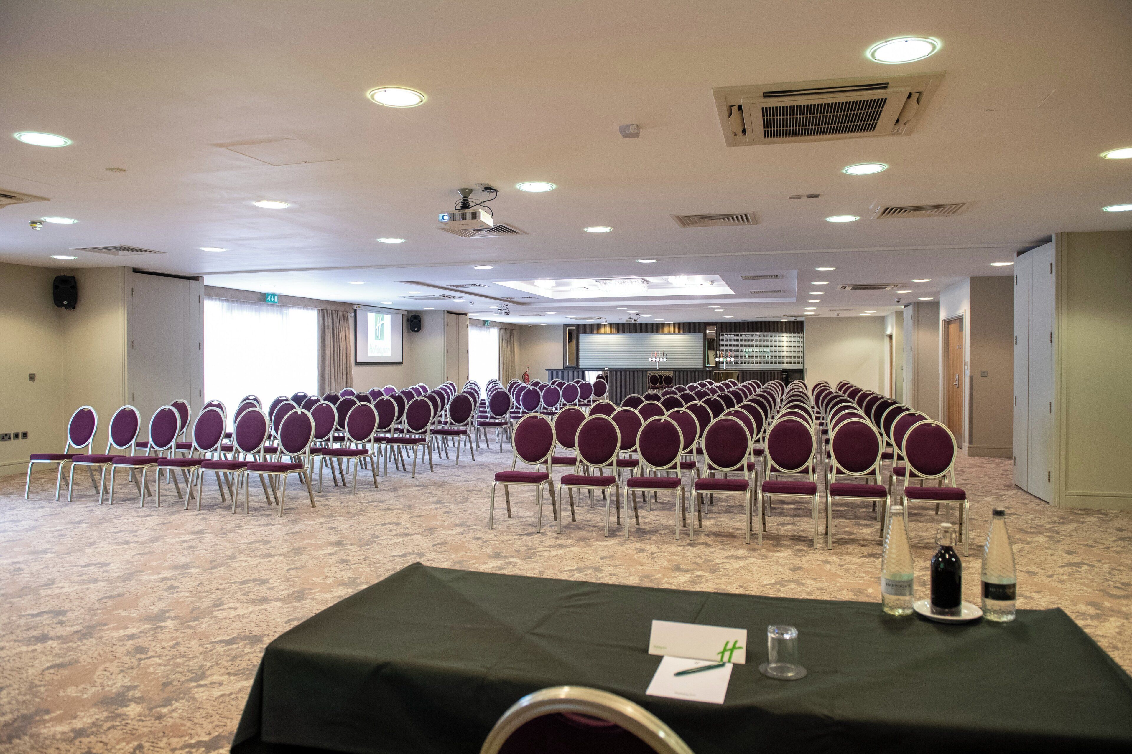 A conference room with rows of chairs, a projector, and a table with water bottles and a notepad.