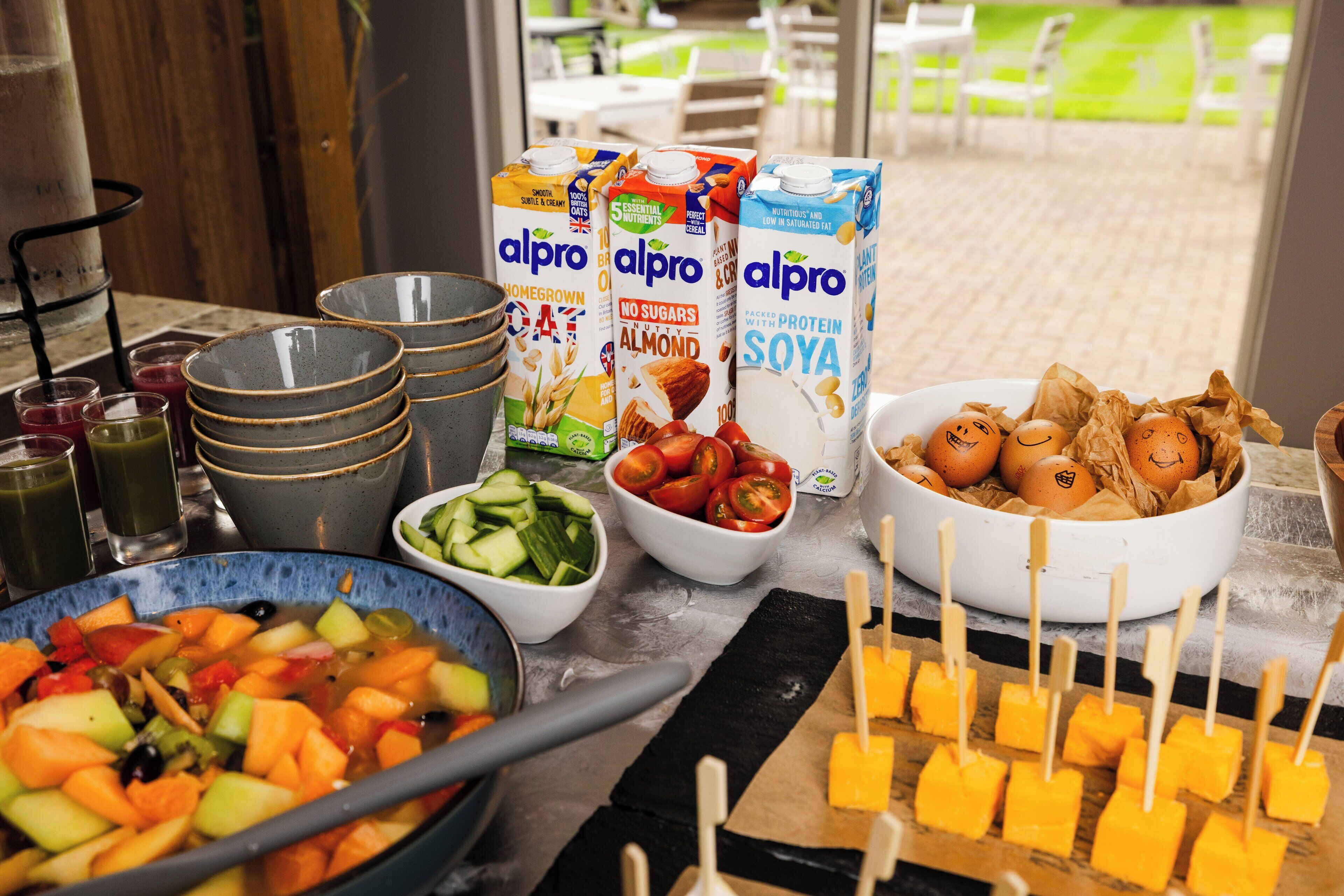 Une table de buffet proposant divers aliments, notamment un saladier de fruits, un saladier de légumes et plusieurs briques de lait d’amande et de lait de soja.