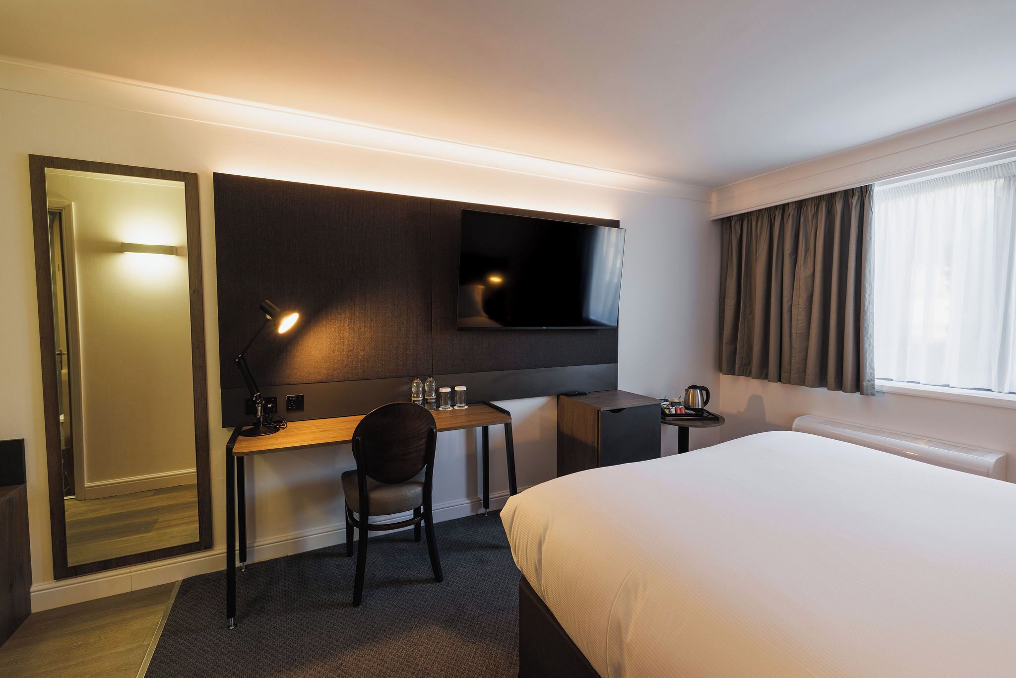 A hotel room with a bed, a desk with a chair, a TV mounted on the wall, and a window with curtains.