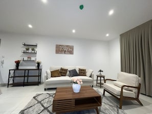 Uma sala de estar moderna com um sofá, uma mesa de centro e uma cadeira. Há um quadro na parede e uma planta na mesinha lateral.