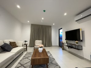 Uma sala de estar moderna com sofá, mesa de centro, TV fixada na parede e uma grande janela com cortinas.