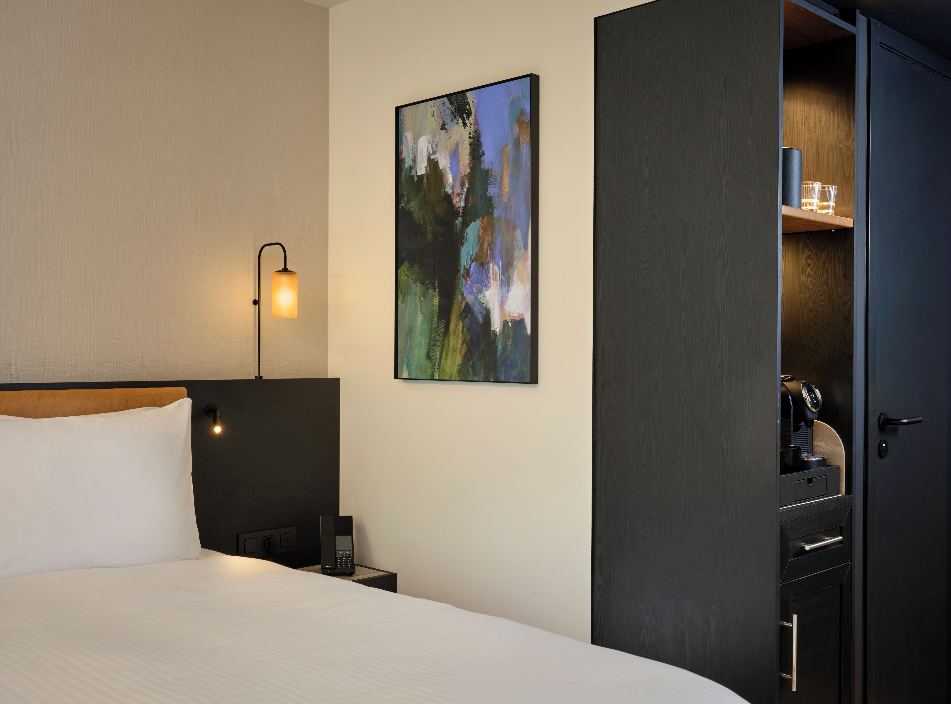 A hotel room with a bed, a bedside table with a lamp, a painting on the wall, and a wardrobe with a coffee machine inside.
