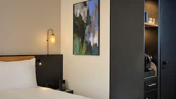 A hotel room with a bed, a bedside table with a lamp, a painting on the wall, and a wardrobe with a coffee machine inside.