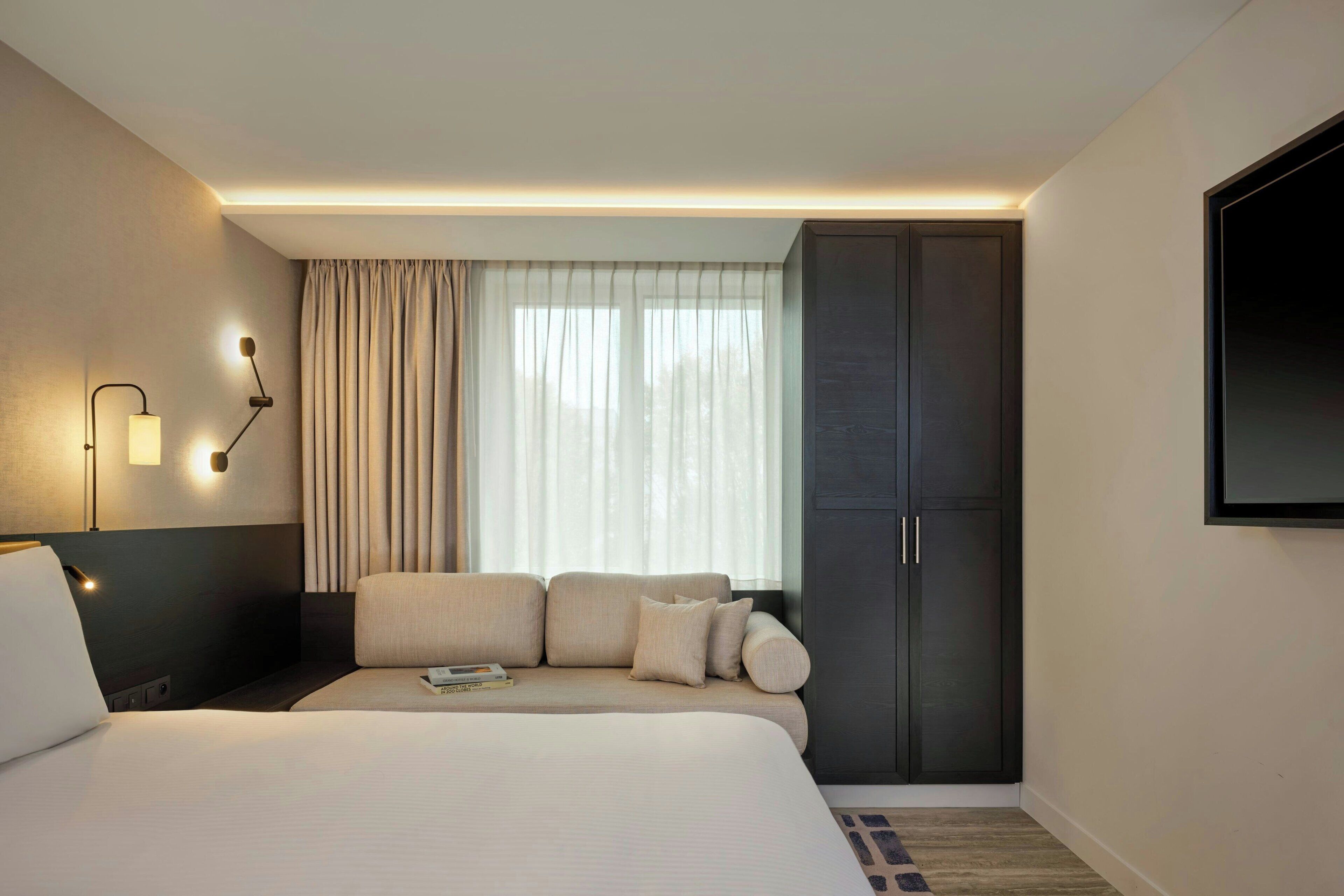A hotel room with a bed, a sofa, a TV, a wardrobe, and a window with curtains.