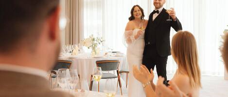 A wedding reception with guests seated at round tables, a couple in wedding attire, and a man in a suit holding a glass.
