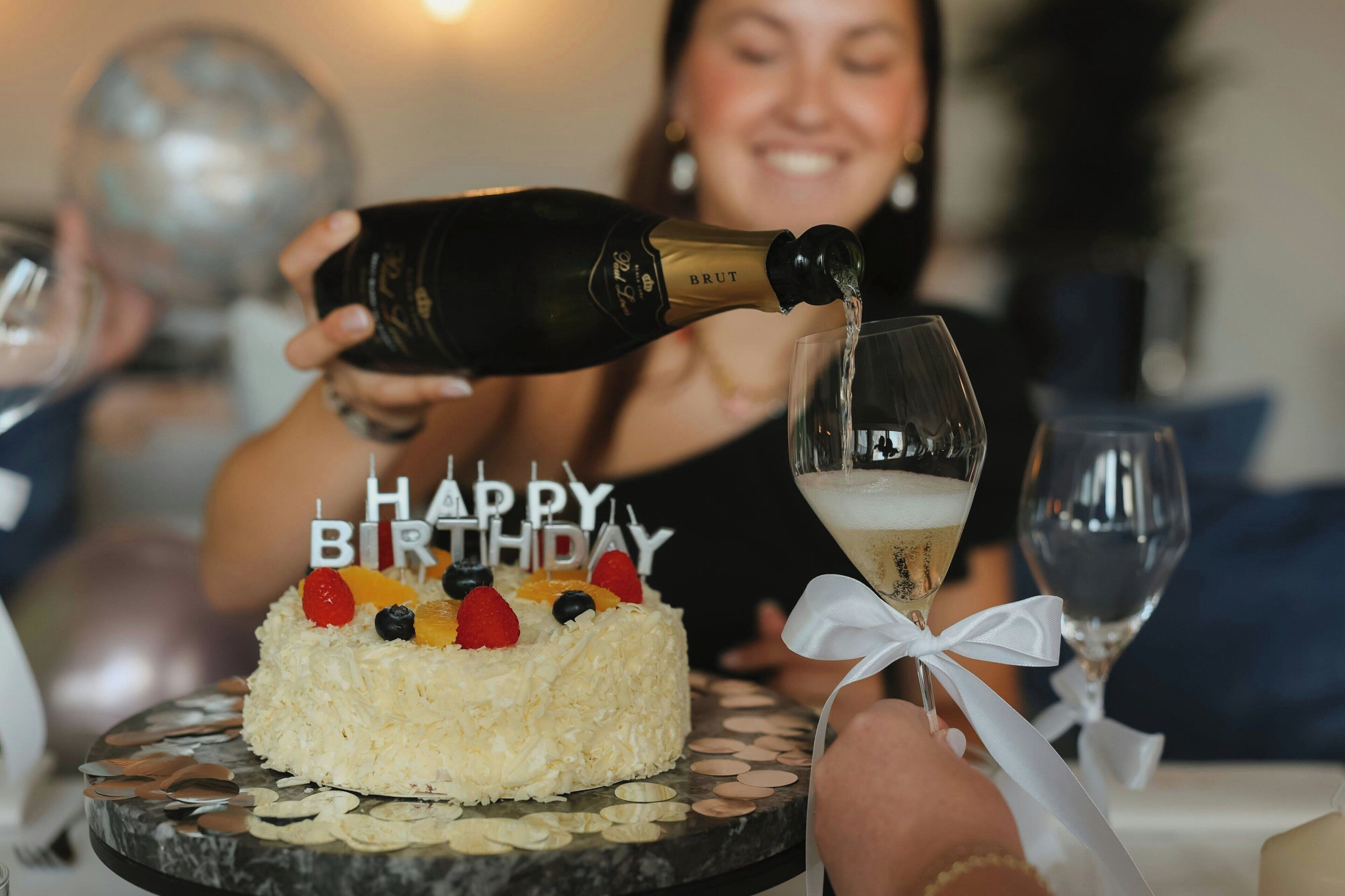 A birthday cake with 'HAPPY BIRTHDAY' candles, a bottle of champagne being poured into a glass, and a person holding a glass.