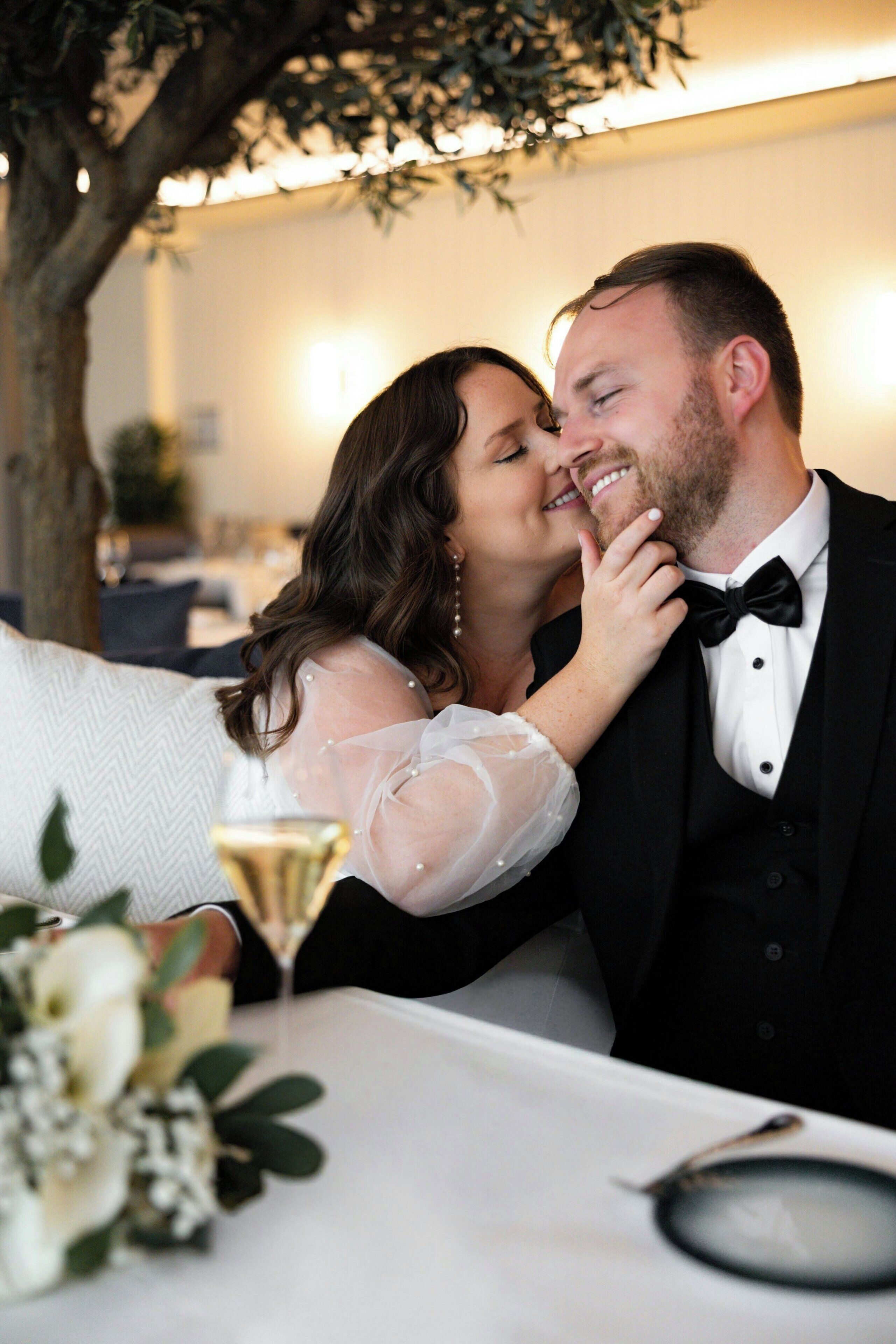 Una pareja sentada a una mesa con mantel blanco, una copa de champán y un arreglo floral.