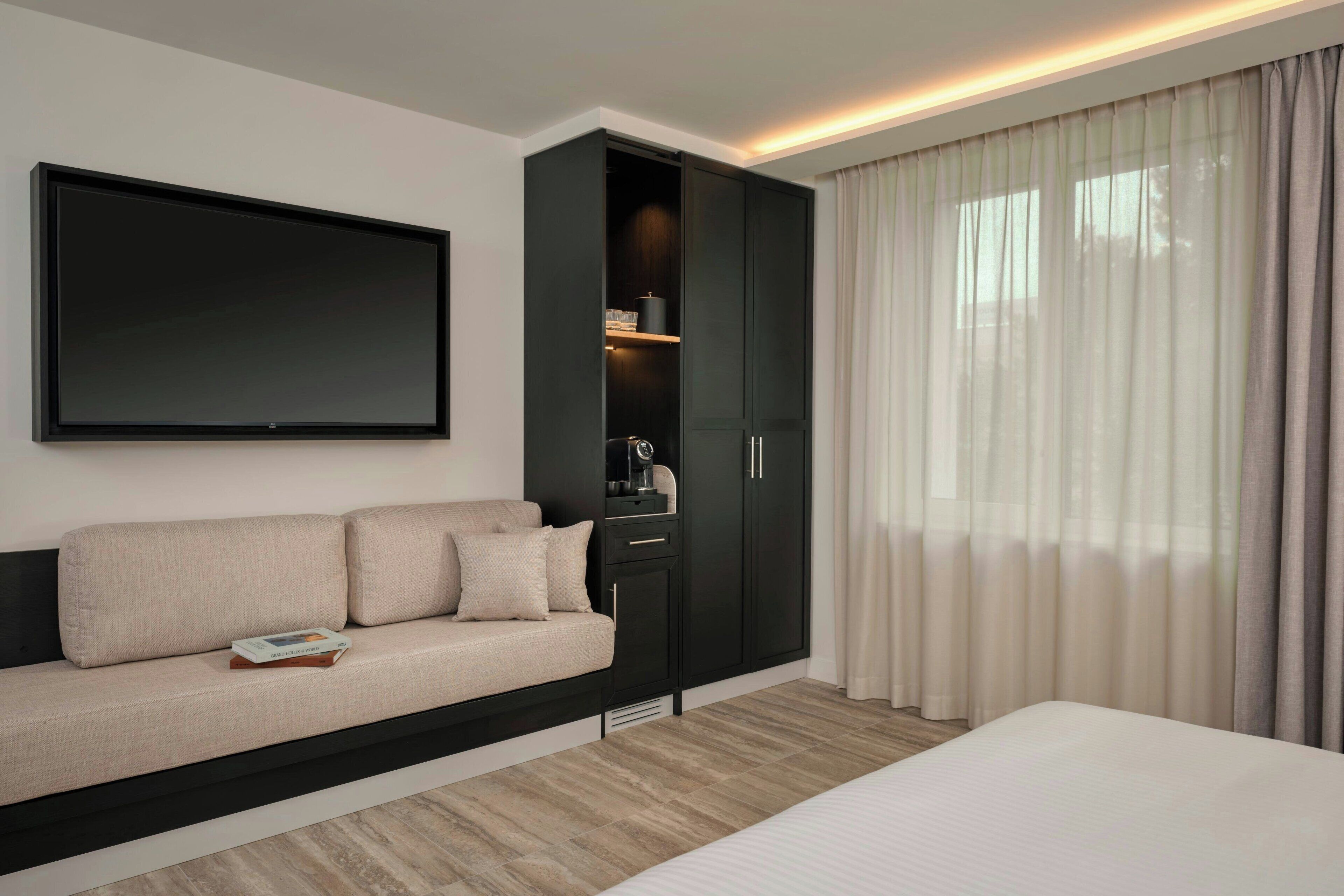 Una habitación de hotel moderna con sofá, televisión de pantalla plana, armario y un gran ventanal con cortinas.