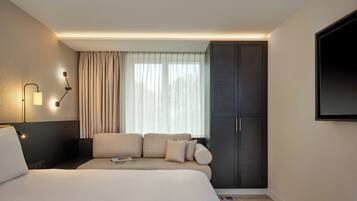 Une chambre d’hôtel moderne avec un grand lit, une armoire sombre, un téléviseur à écran plat fixé au mur et une fenêtre ornée de rideaux légers.
